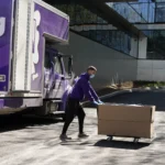 A mover in a purple hoodie pushing a large box on a cart past a Perfect Moving truck