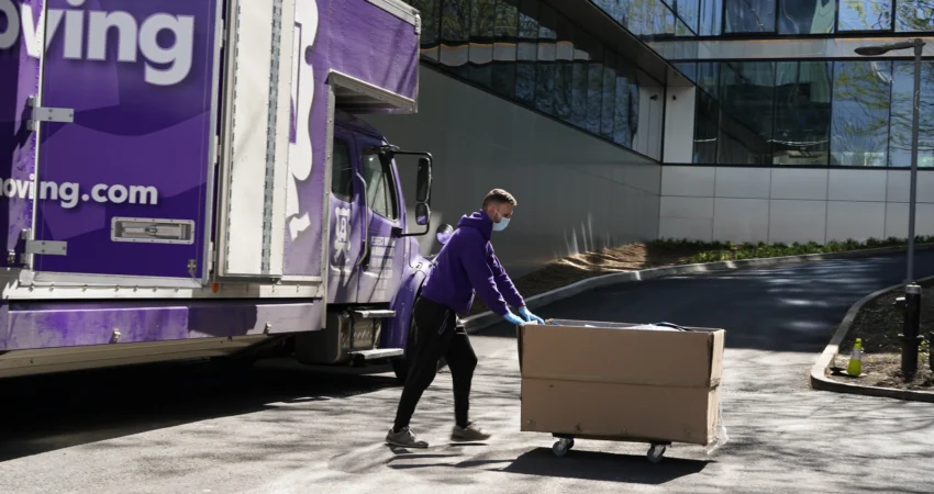 A mover in a purple hoodie pushing a large box on a cart past a Perfect Moving truck